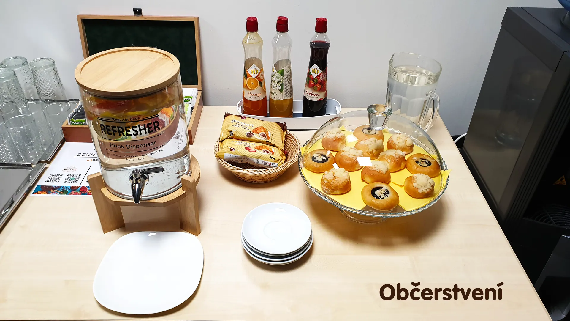 Table with sweet pastries, fruit syrups, a water dispenser, and glasses set up for training participants.